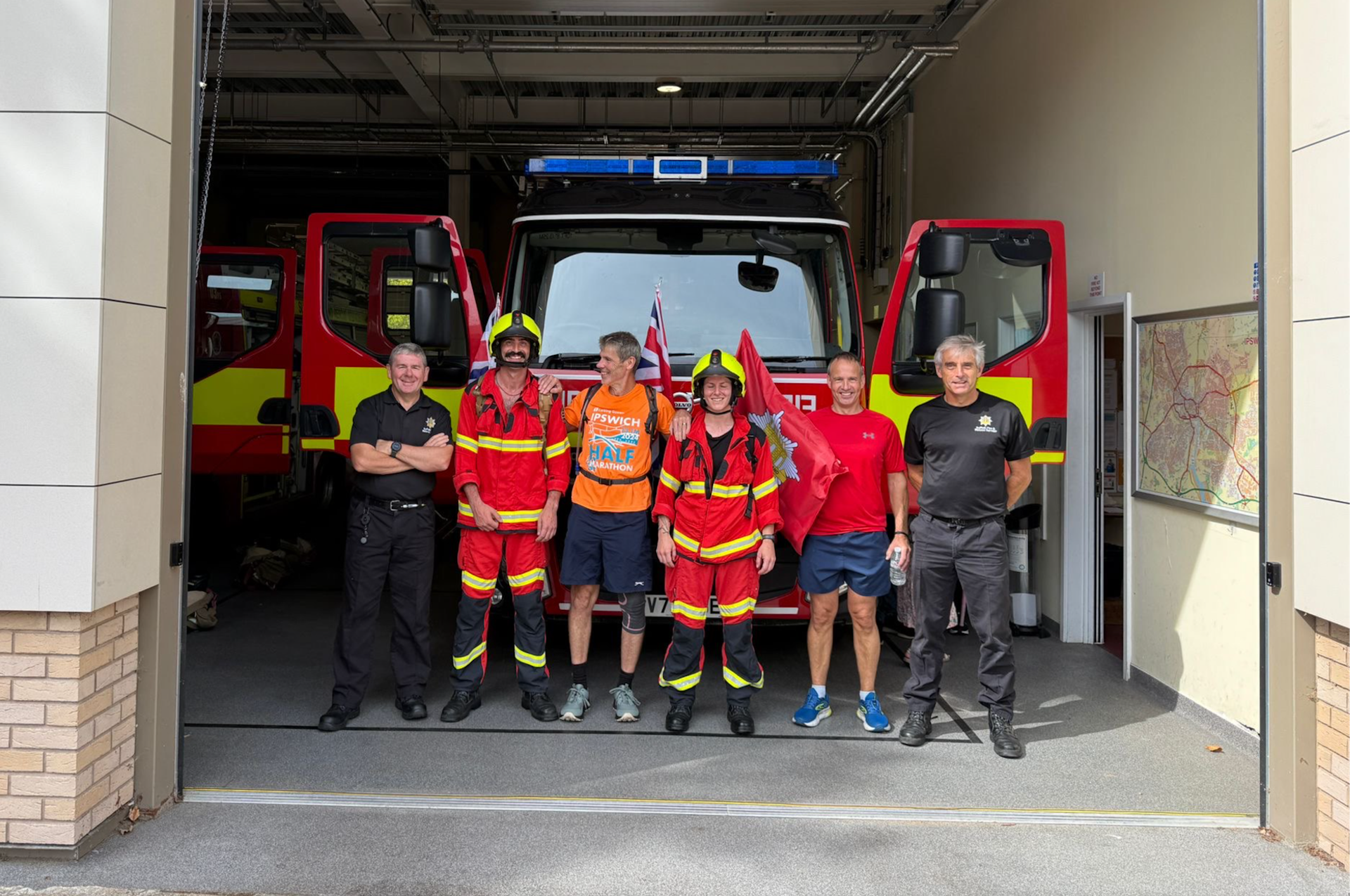 Six Suffolk firefighters in kit and running gear stand in front of two fire engines, preparing to carry a stretcher in the Ipswich Half Marathon